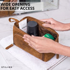 Leren toilettas in tan/donkerbruin, wijde opening met zichtbare binnenvakken, handen plaatsen toiletartikelen, STILORD-logo zichtbaar